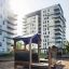 Legehus med sandkasse Classic Natur placeret i en bypark med moderne bygninger i baggrunden.